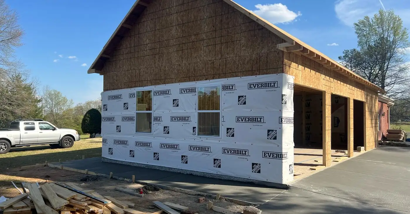 Framed garage building wrapped in house wrap beside a paved drive