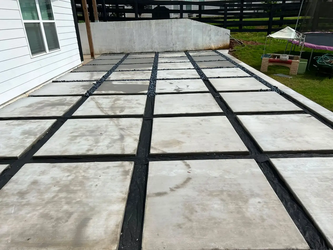 Large concrete slab patio set in a grid with black river-rock joints beside a house